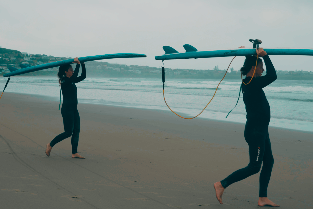 Two people in surf wetsuits carrying surfboards on their heads